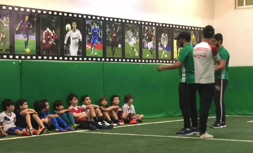 Enfants assis au bord du terrain de foot indoor devant un mur de légendes du football