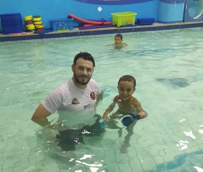 Un coach KidiClub tient un tout-petit souriant dans la piscine, moment de confiance pendant un cours de bébé nageur